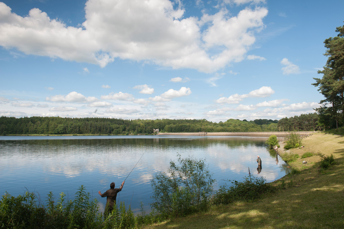 Fishermen stood at the edge of a reservoir with a grass banking behind them and a large expanse of woodland and dam in the distance at the far side of the reservoir