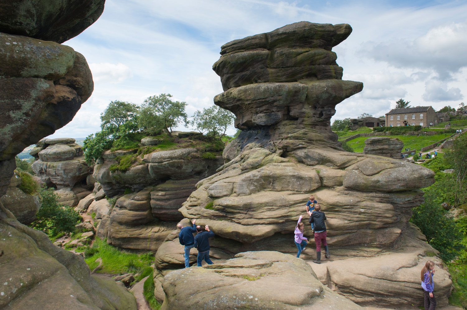 A series of rock formations at Brimham rocks with adults and children standing on them and a large building in the background to the right