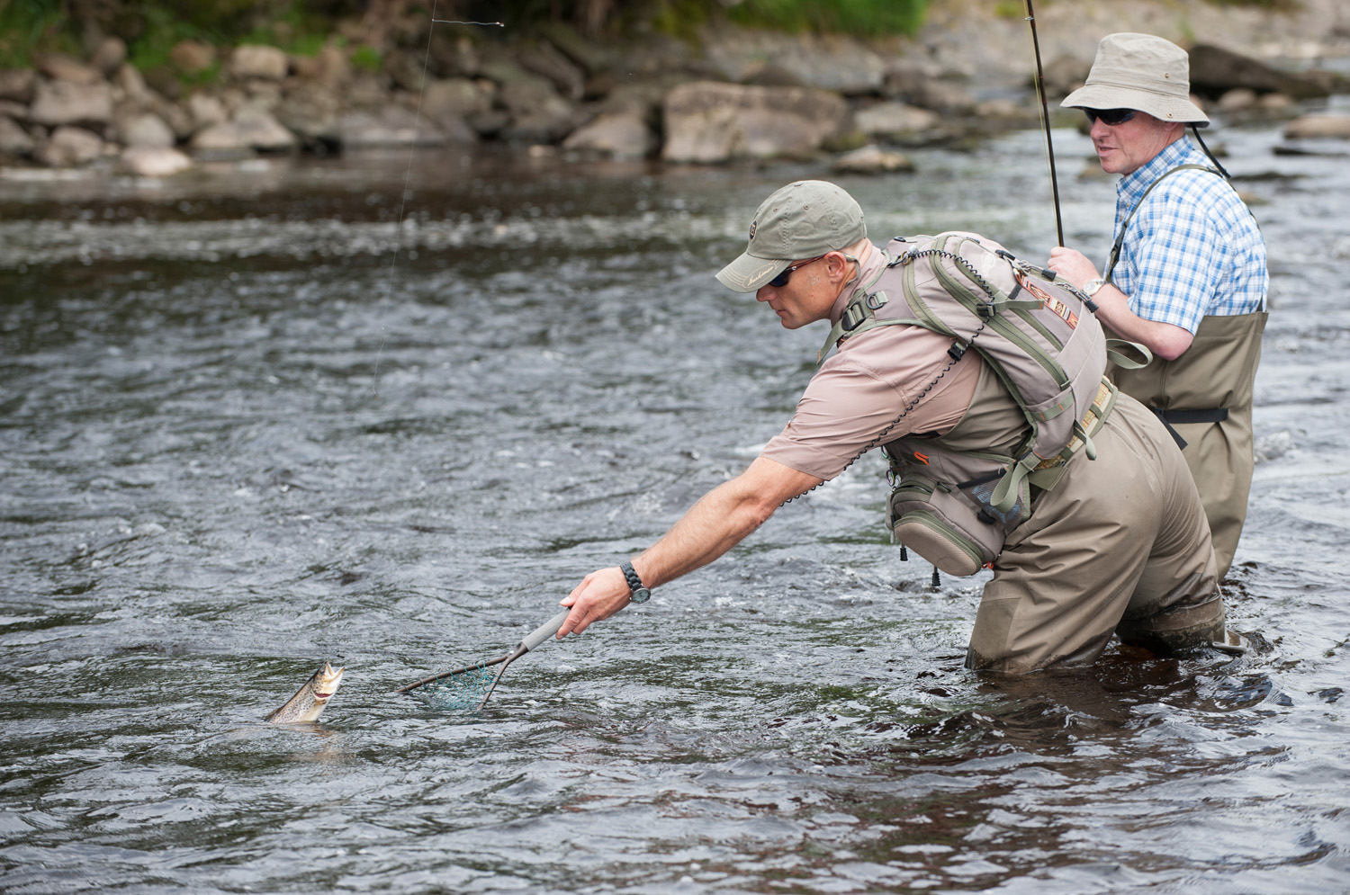 Two fishermen stood in a river, one holding a net in the water at arms length with a fish popping out of the water