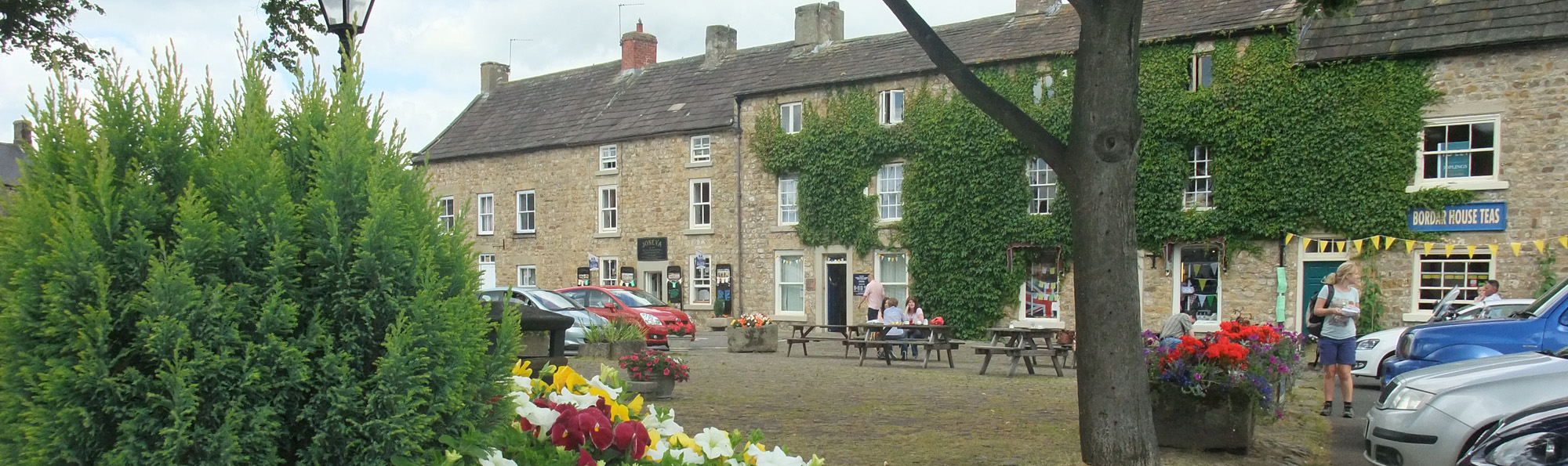 A row of three storey buildings with some cars parked outside , people sitting and walking and some flower filled planters
