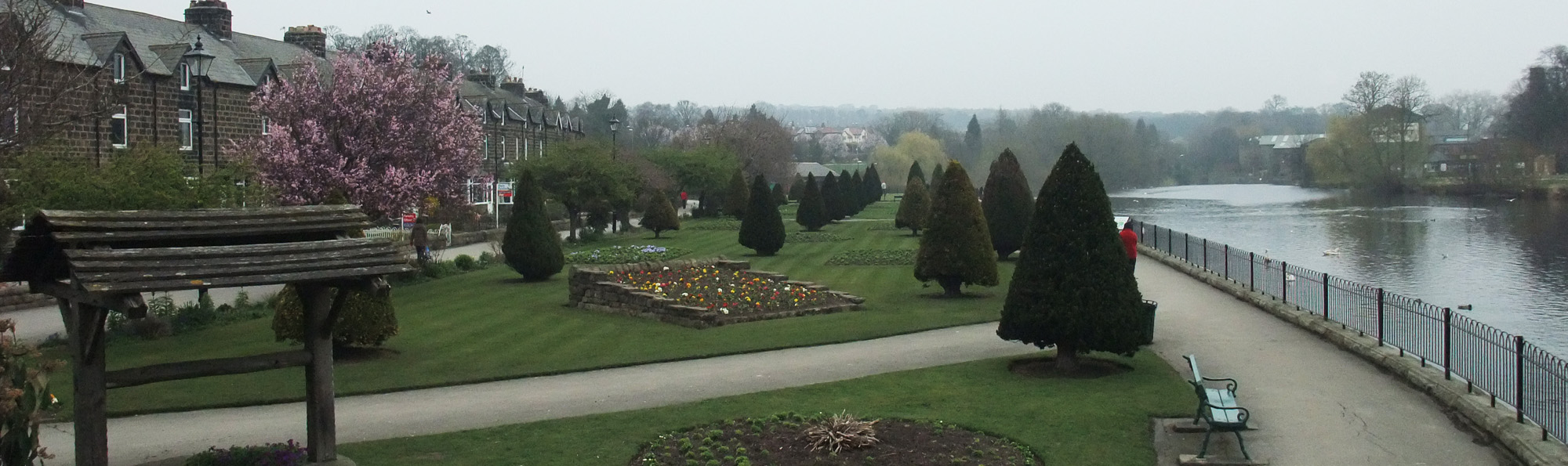 A row of houses and an expanse of grass, flower beds and triangular shaped trees with a fenced off river to the right