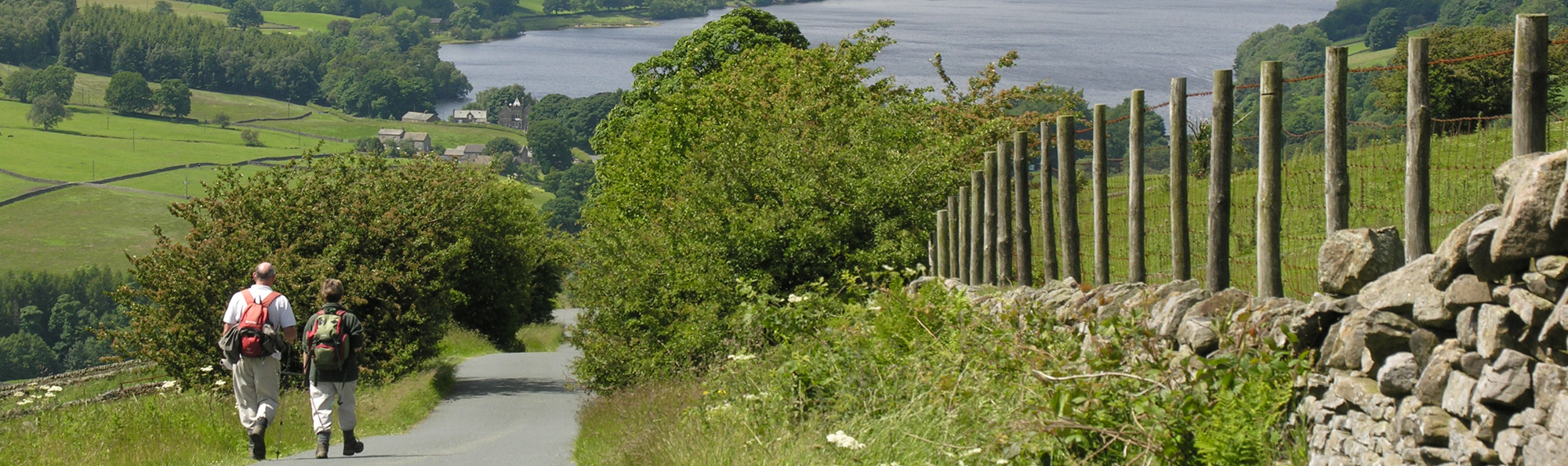 Two walkers wearing backpacks walking down a country lane with Gouthwaite reservoir beyond