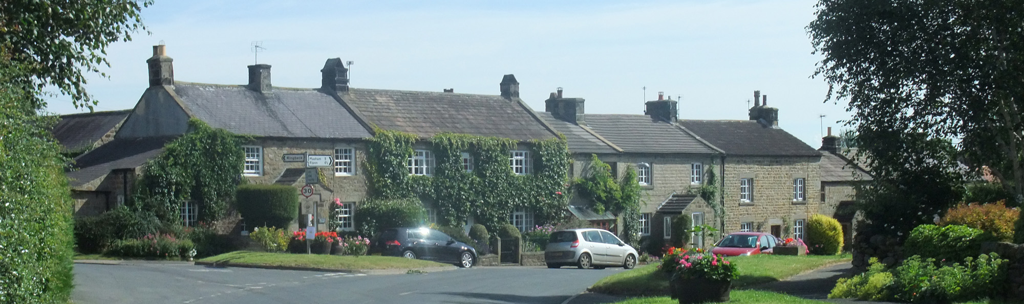A row of houses, with cars parked outside, and a signpost at the side of a road in the village of Kirkby Malzeard