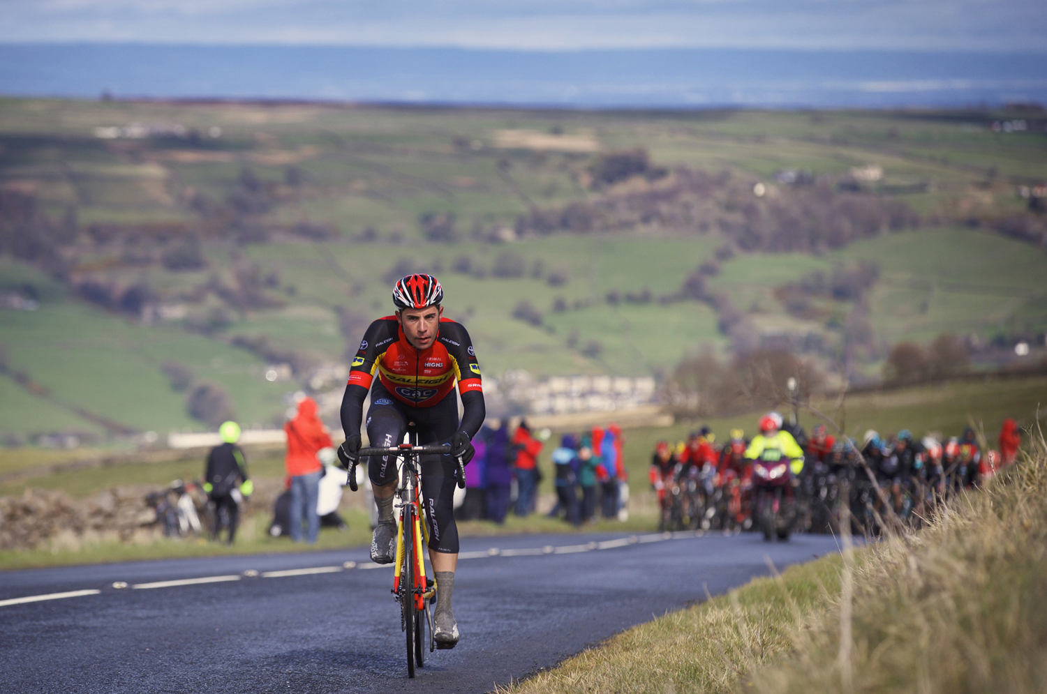 A close up of a cyclist on a road with people, and a large group of cyclists behind