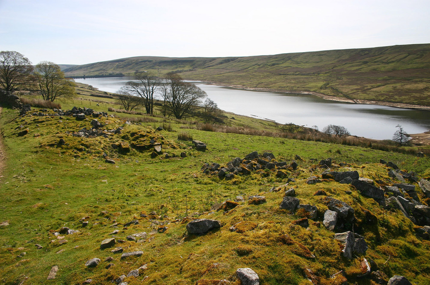 Looking down across Scar House reservoir with the remains of Lodge village and a few bare trees in the foreground and Scar House dam in the distance