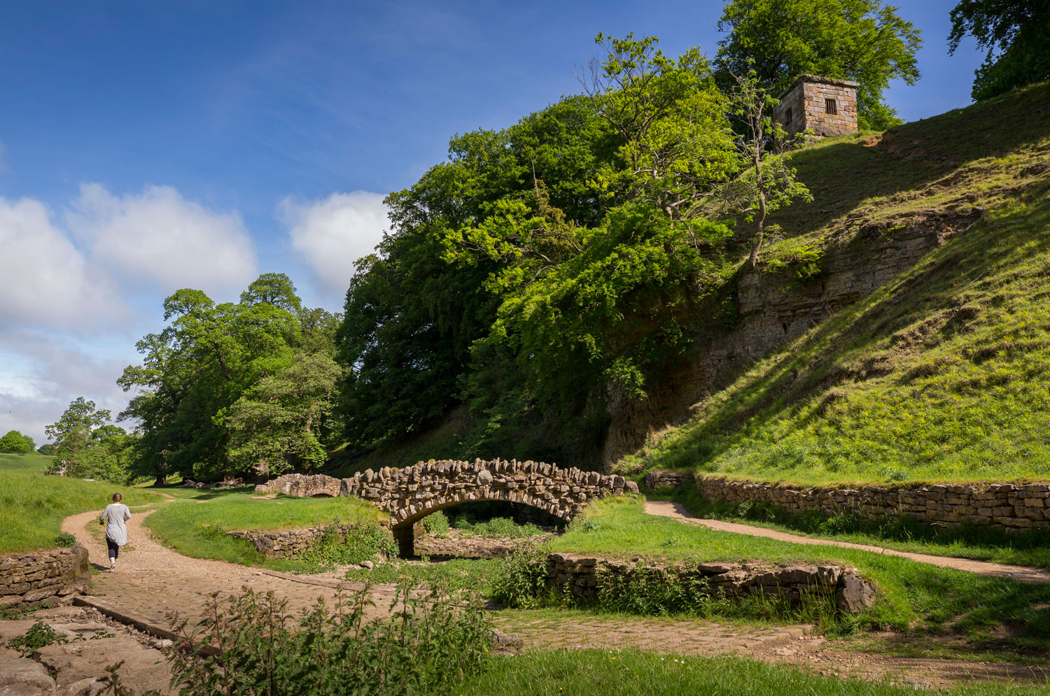 Two stone bridges in the Seven Bridges Valley with someone walking on the path alongside and trees and a building on the hillside above