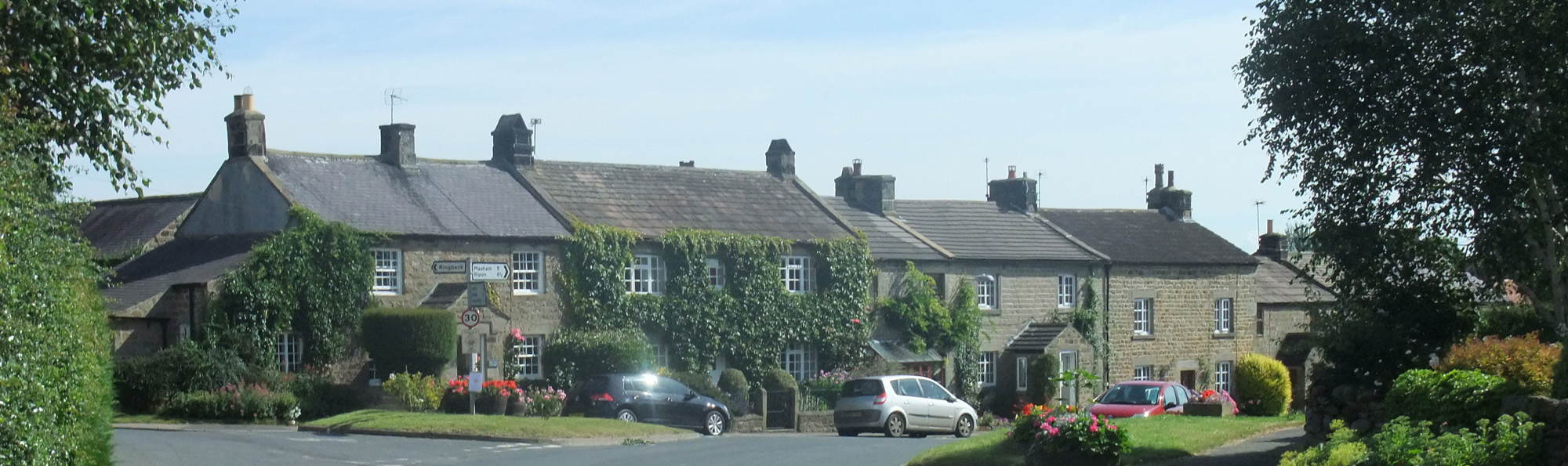 A row of houses, with cars parked outside, and a signpost at the side of a road in the village of Kirkby Malzeard