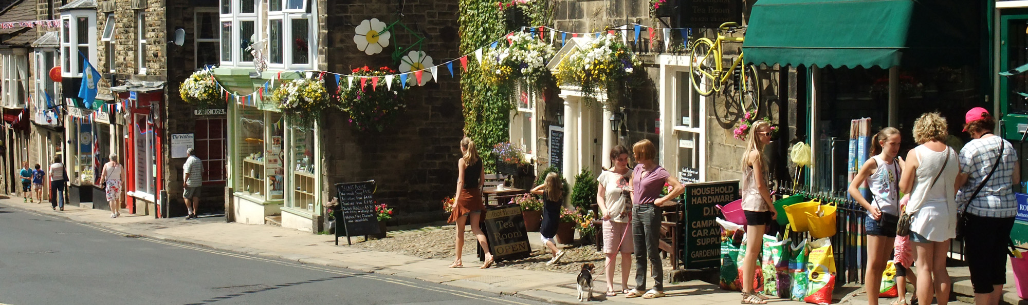 People on the pavement outside shops, with bunting and flowers on display