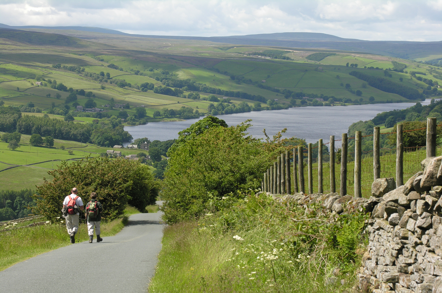 Two walkers wearing backpacks walking down a country lane with Gouthwaite reservoir, green fields and hillsides beyond
