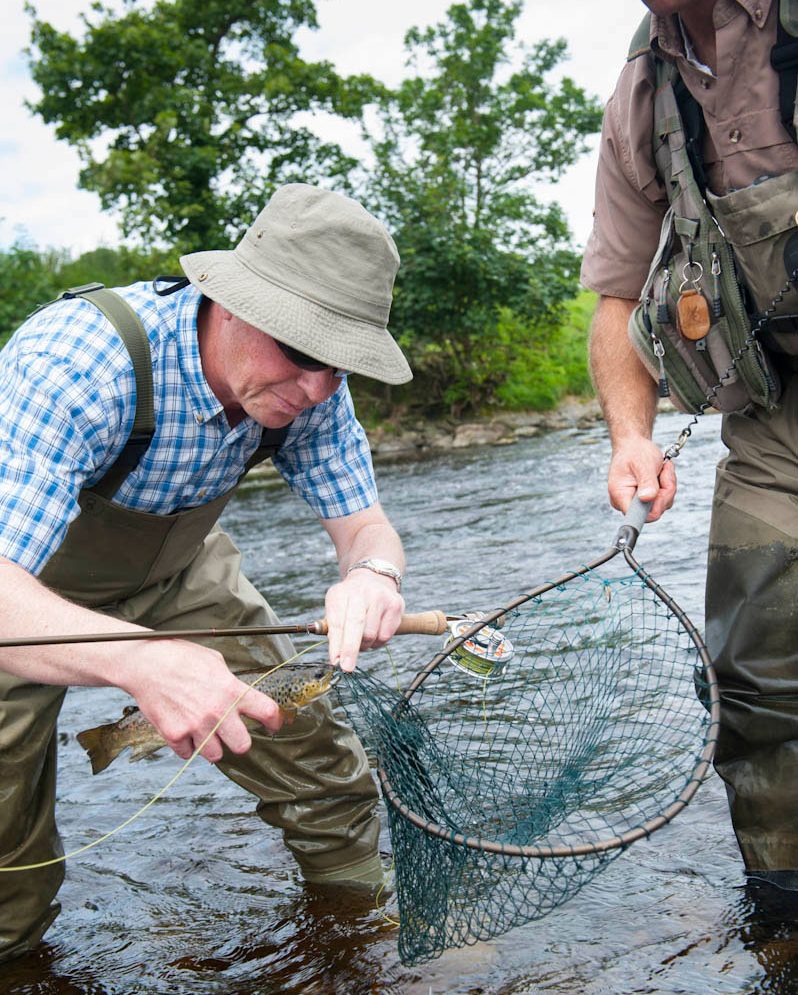 Two fisherman stood in a river, one holding a fish in one hand and his rod in the other, the other holding a net