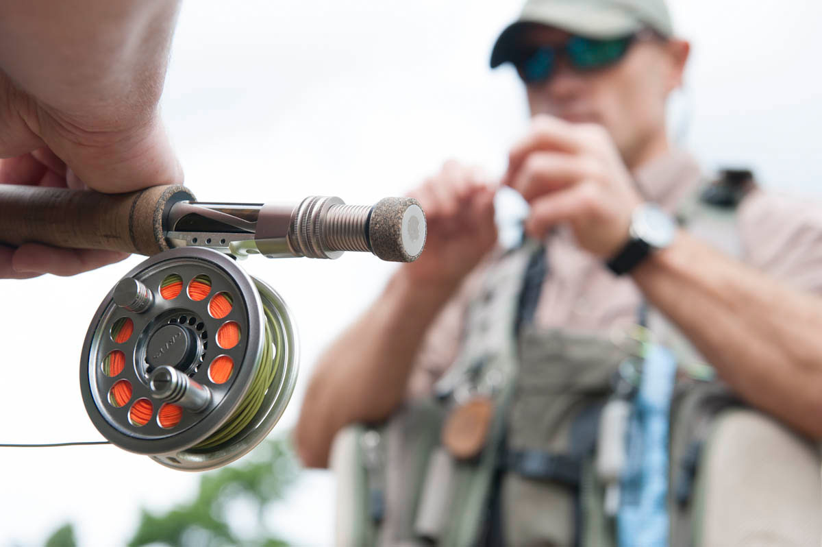 A close up of a hand holding a fishing rod reel with orange thread and a blurred fisherman in the background